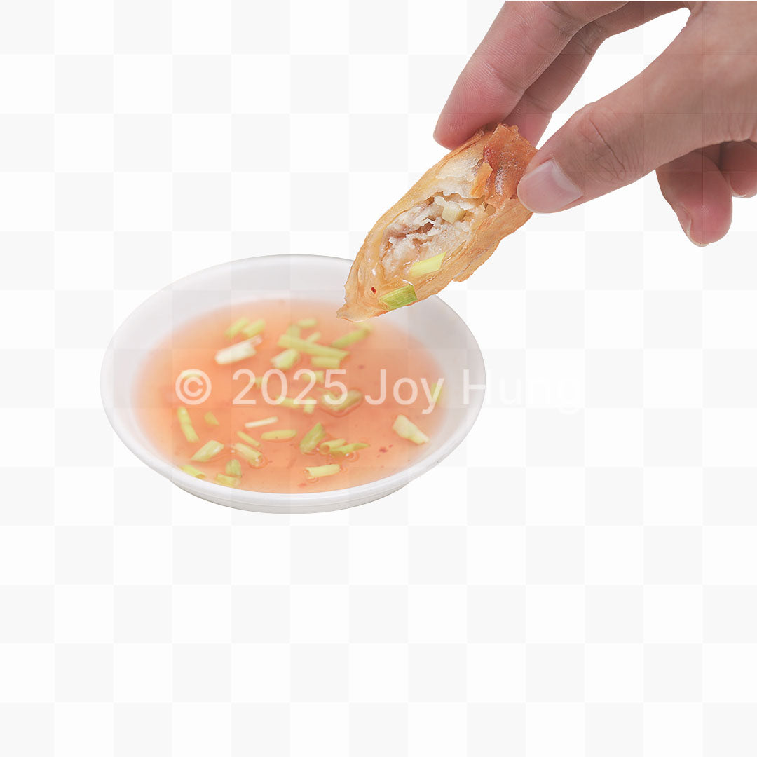 Hand dipping a fried lumpia into a small bowl of sauce with green onions on a white background.