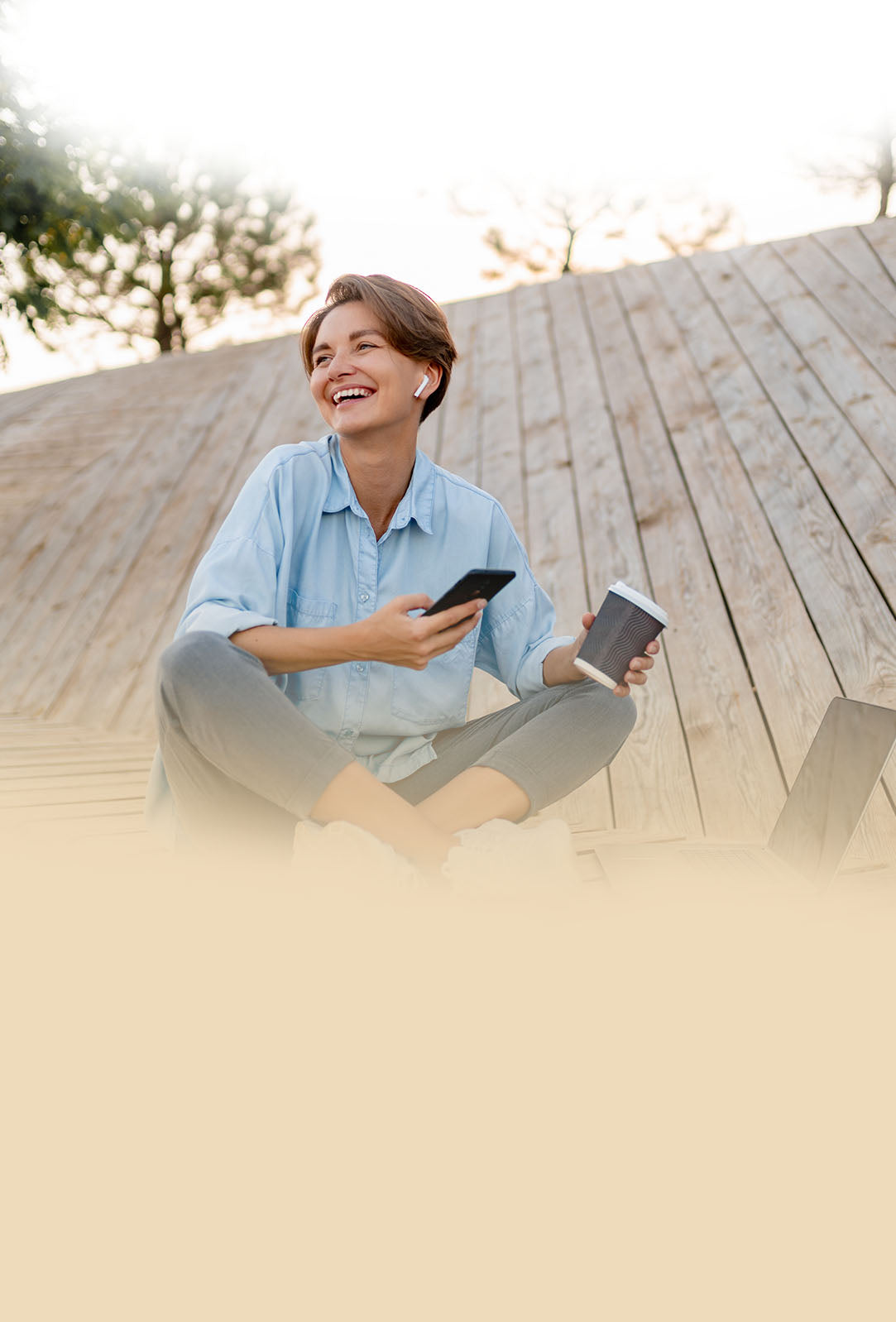 Woman sitting on a wooden deck holding a phone and a coffee cup, smiling.