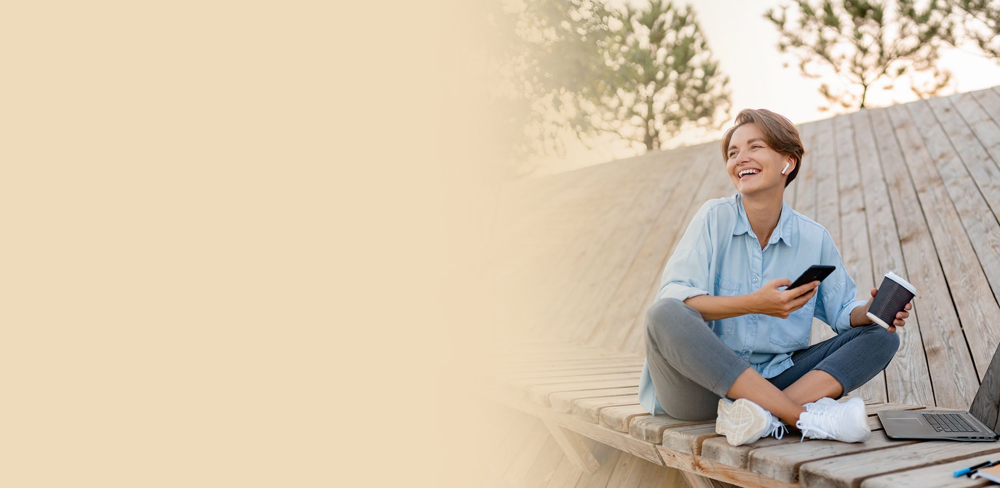 Woman sitting on a wooden bench outdoors, using a smartphone and laptop.