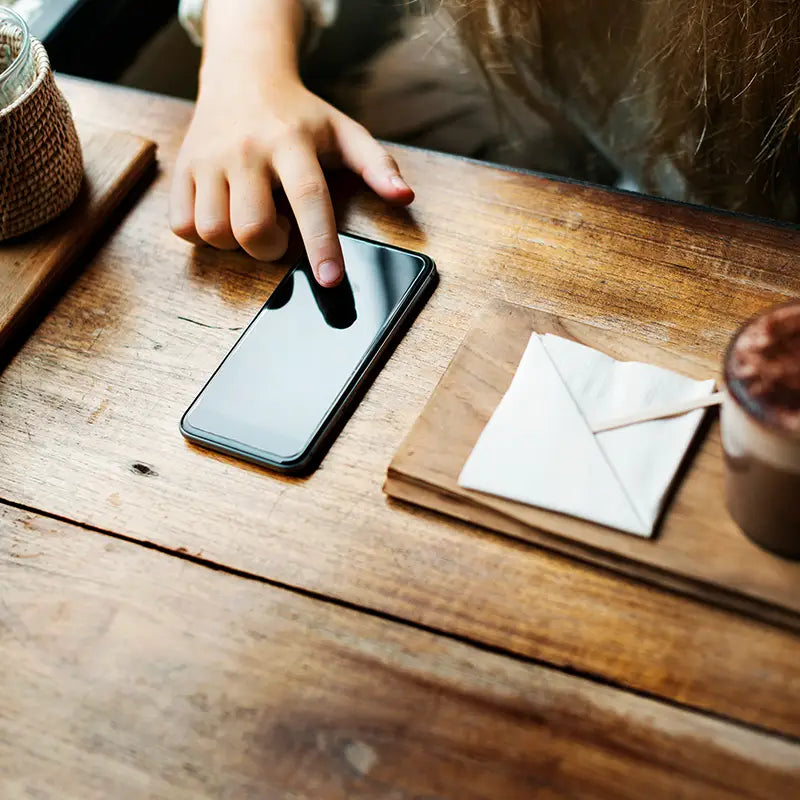 Person using a smartphone on a wooden table with a cup of coffee and a napkin.