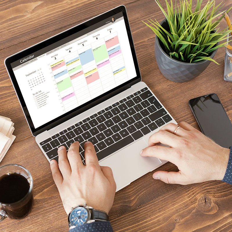 Person using a laptop with a calendar on the screen, sitting at a wooden desk with a plant and phone.