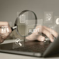 Person using a magnifying glass over a laptop with audit-related icons on a blurred background