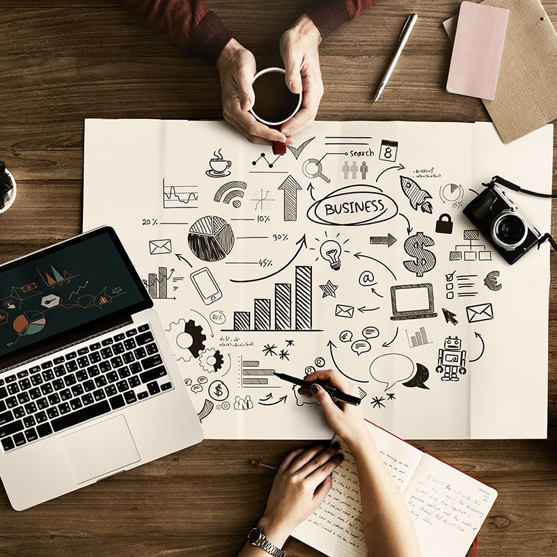 Two people working at a desk with a laptop, coffee, and various business-related illustrations on a large sheet of paper.