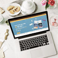 Laptop on a table with a website open displaying supplements, surrounded by tea set and cookies.