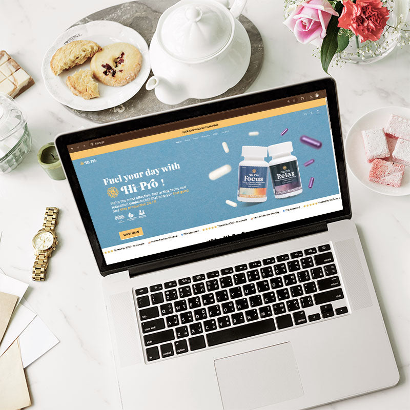 Laptop on a table with a website open displaying supplements, surrounded by tea set and cookies.